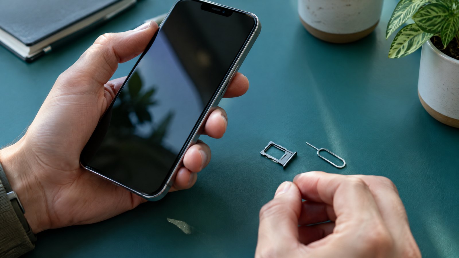 A person holds an unbranded smartphone and a tiny removable SIM beside a SIM-eject pin on a desk