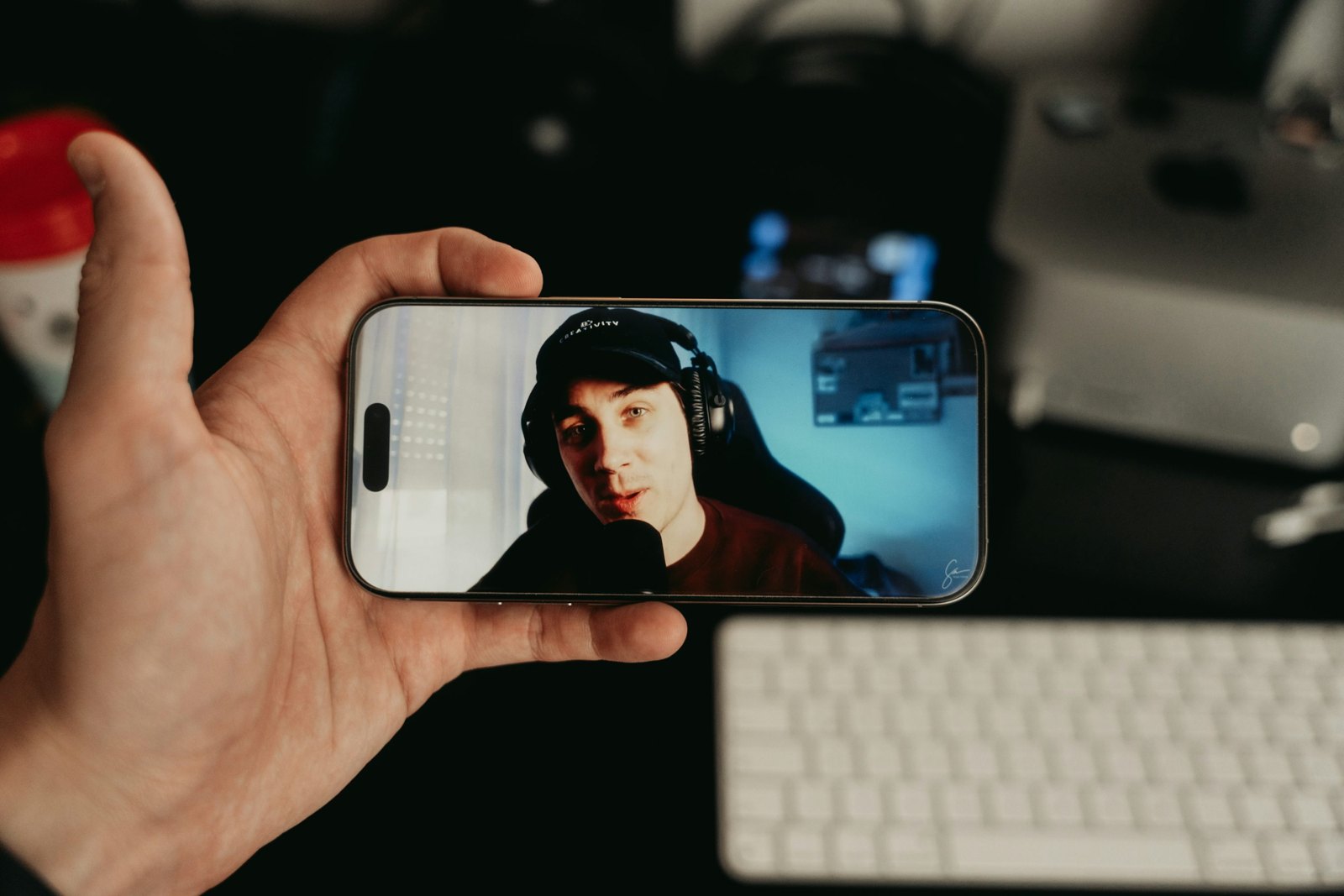 A hand holding a phone that is playing a video, with a computer desk blurred in the background.