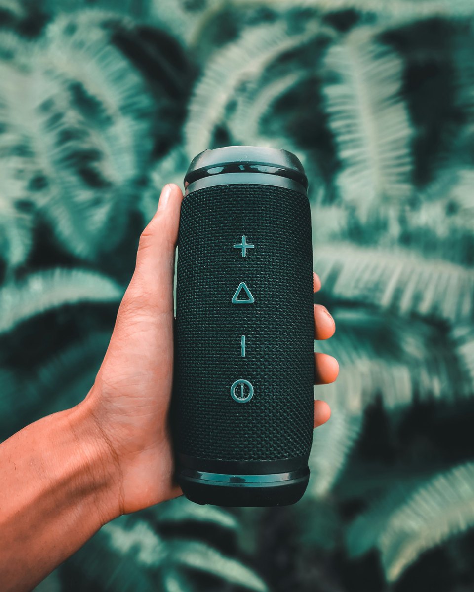 A hand holds a generic black portable Bluetooth speaker outdoors; it is not a CMF Speaker.