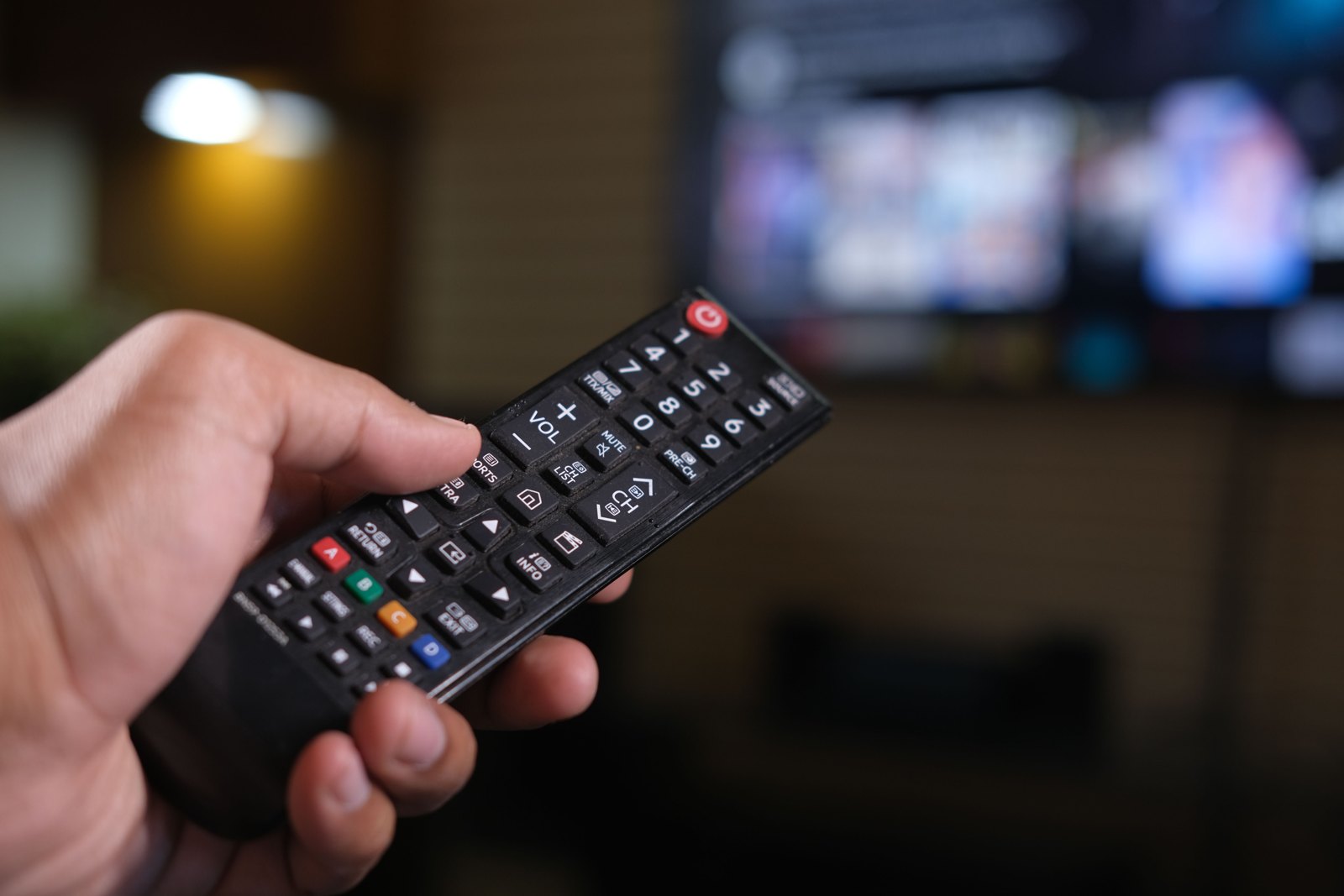 Hand holding a generic television remote in front of a blurred television screen