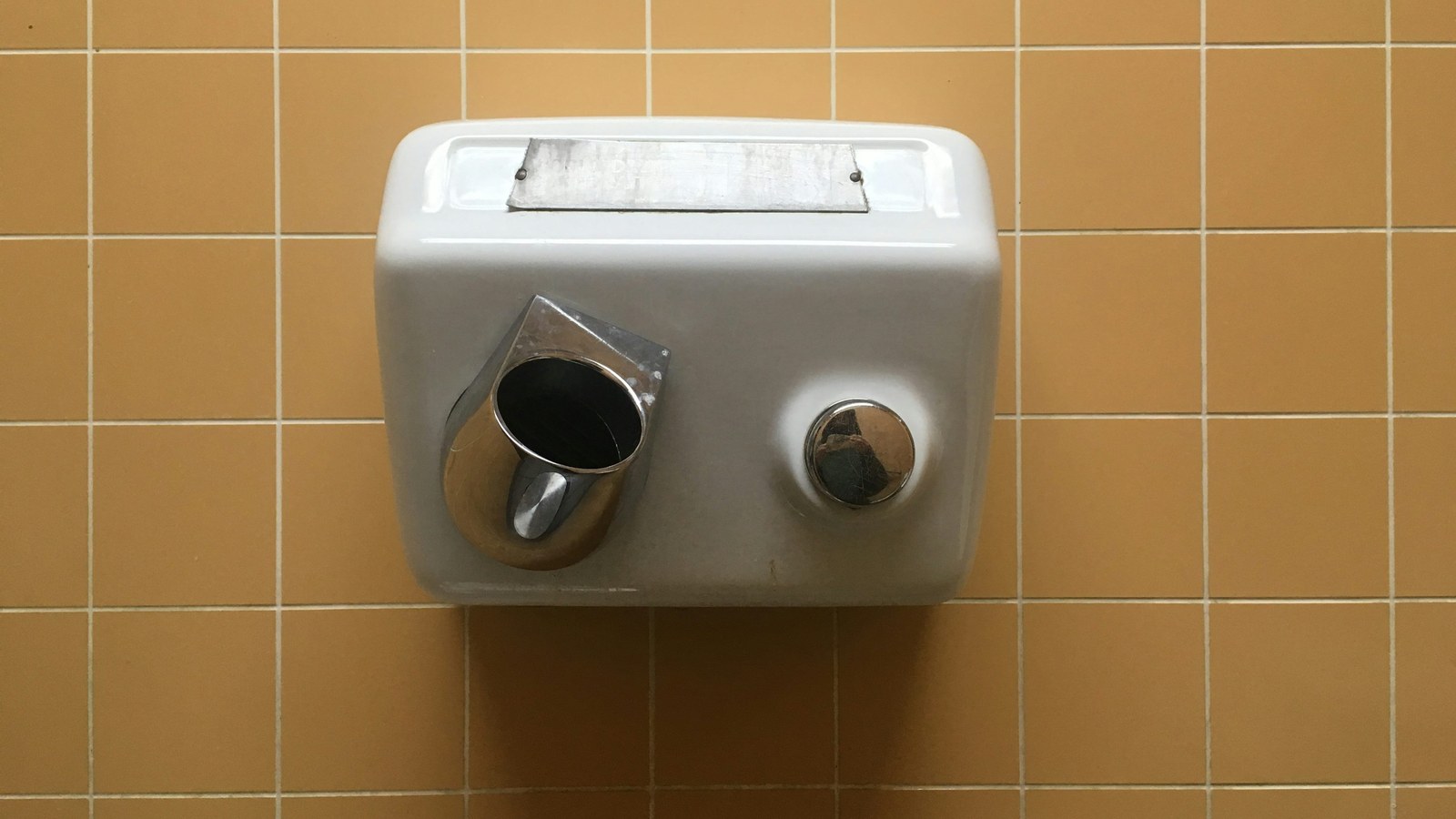 Conventional metal push-button hand dryer mounted on yellow bathroom tiles