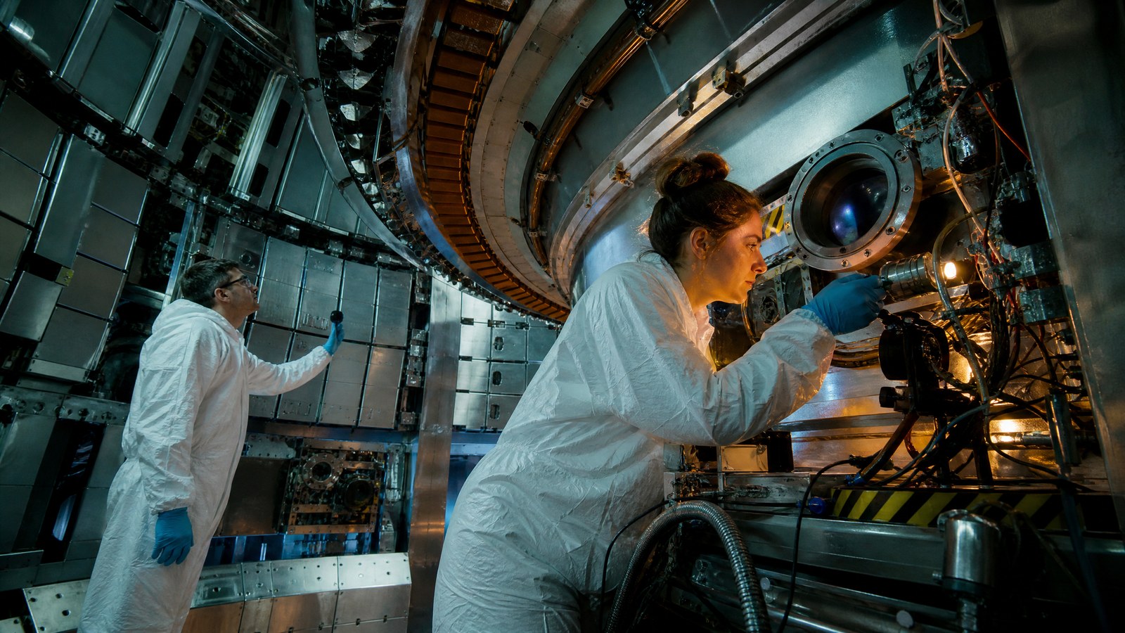 Two plasma physicists inspect diagnostic ports and cables around a compact experimental fusion device.