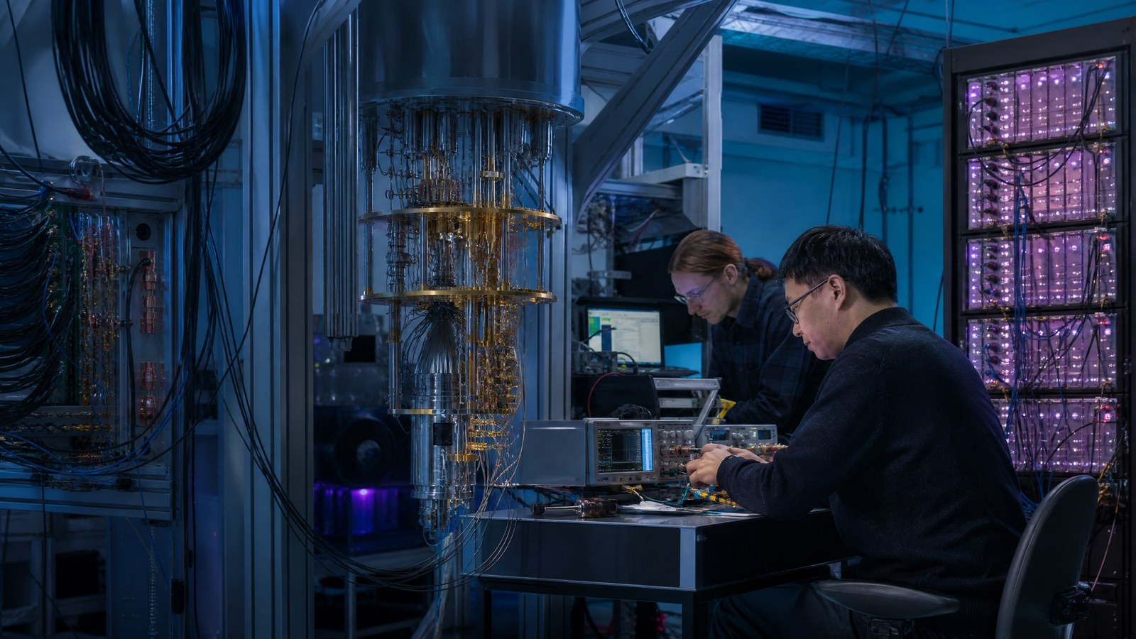 Two quantum computing researchers calibrate cable connections beside a cryogenic control rack in a blue-lit laboratory.